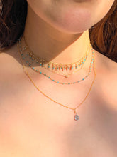 Load image into Gallery viewer, A close-up of a person's neck wearing multiple layered gold necklaces, including one with small turquoise-blue beads and one with a clear teardrop crystal pendant, in warm natural sunlight.