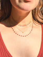 Load image into Gallery viewer, Close-up of a woman's neck and upper chest wearing three layered gold necklaces, including a beaded necklace with red stones and delicate pendant chains, under warm sunlight.