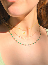 Load image into Gallery viewer, A close-up of a person's neck and chest wearing three layered necklaces: a gold chain with a small green teardrop pendant, a rose gold infinity-style chain, and a beaded chain with green beads.